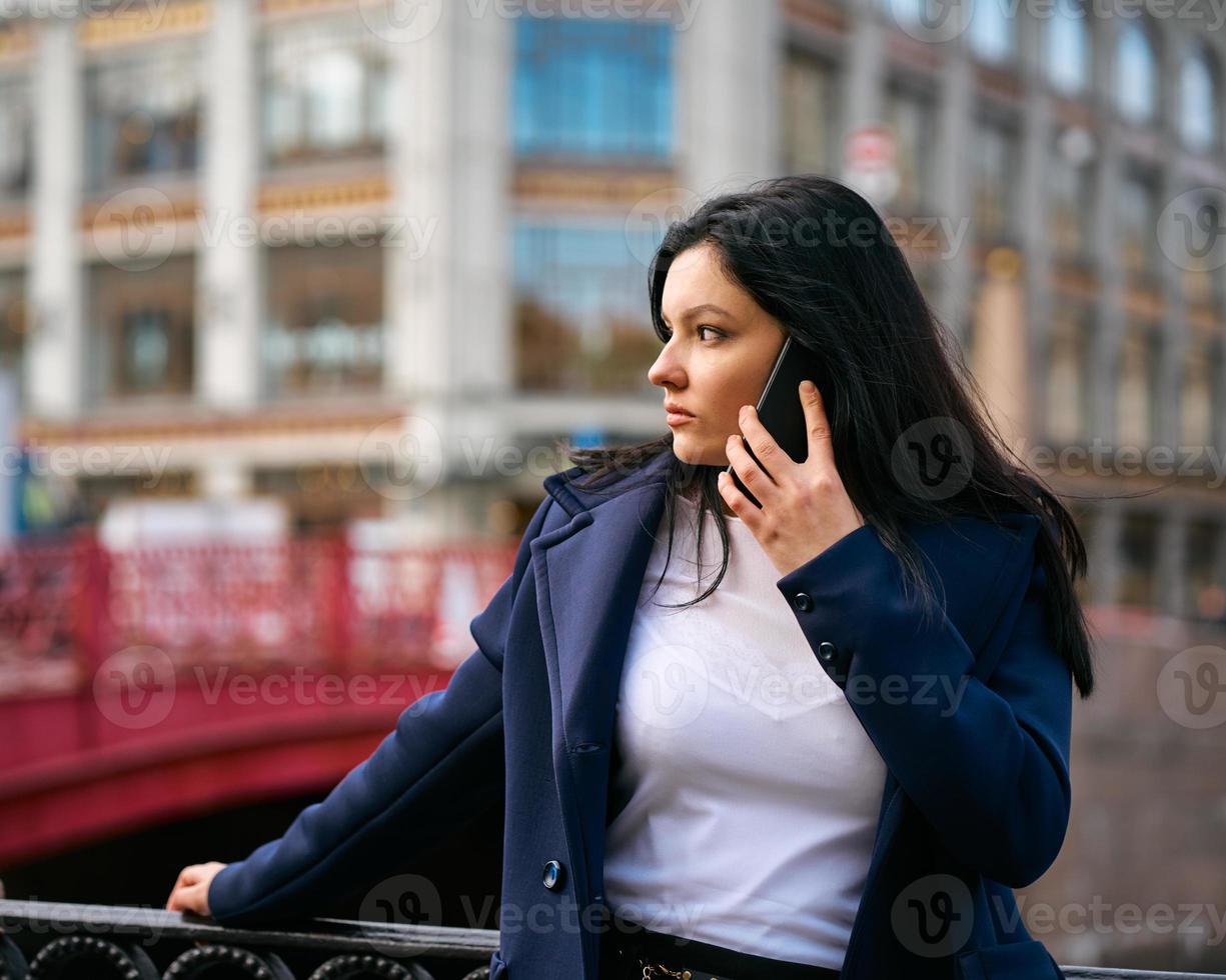 mujer con cabello largo hablando por teléfono. otoño o invierno, chica al aire libre. hermosa morena inteligente en la calle de san petersburgo en el centro de la ciudad cerca del puente, espacio de copia foto