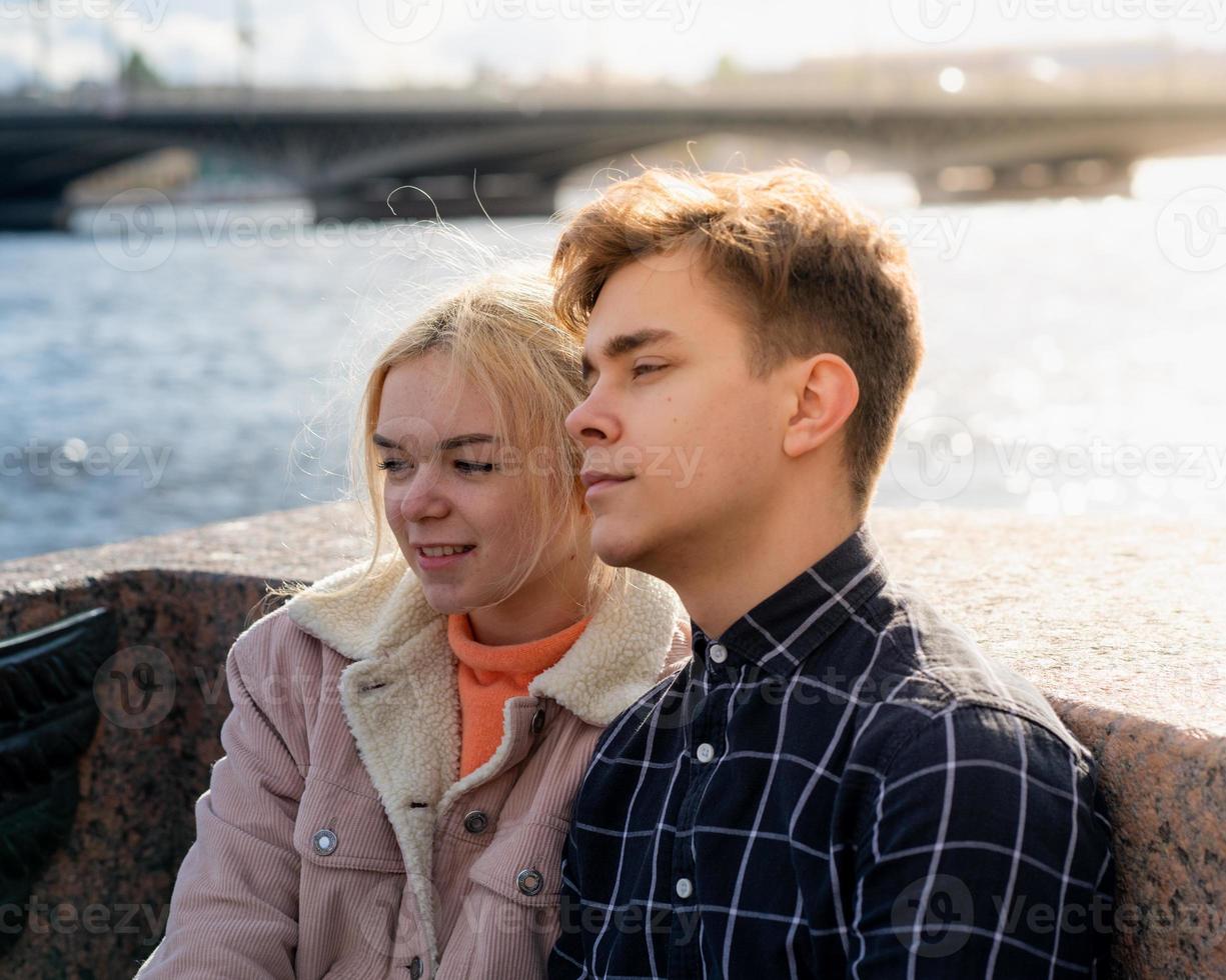 Teenagers in love sit on the waterfront in the city center, resting in autumn sun. Concept the first teenage love, the beginning of relations. A boy and a girl meet on a date, a couple. photo