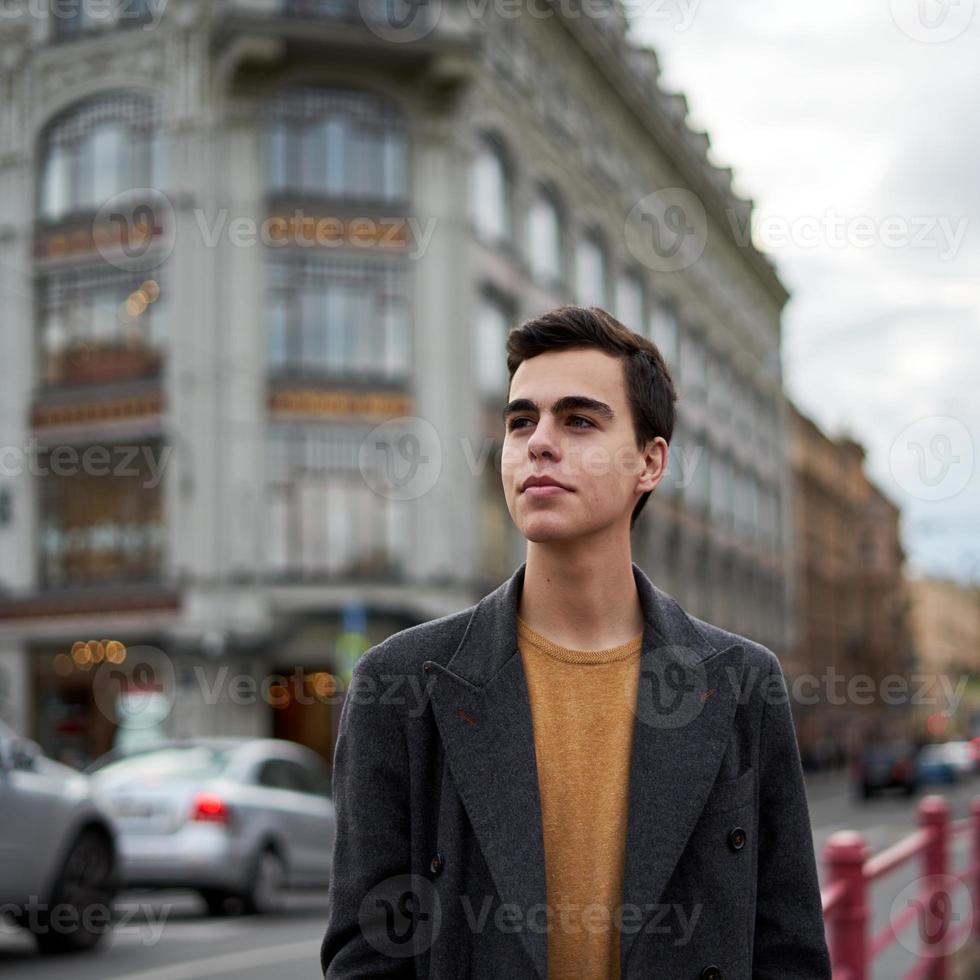 Handsome stylish fashionable man, brunette in elegant gray coat, stands on street in historical center of St. Petersburg. Young man with dark hair, thick eyebrows. photo