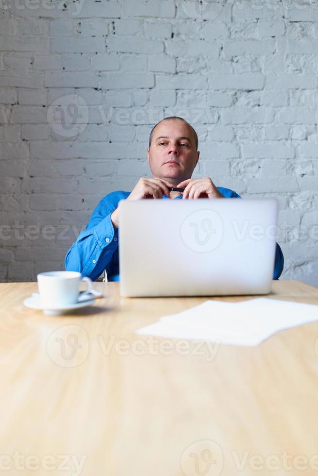 Handsome handsome mature man sitting at table in front of laptop and listening to subordinate. Man with casual clothes in blue shirt in the office in front of window, vertical photo