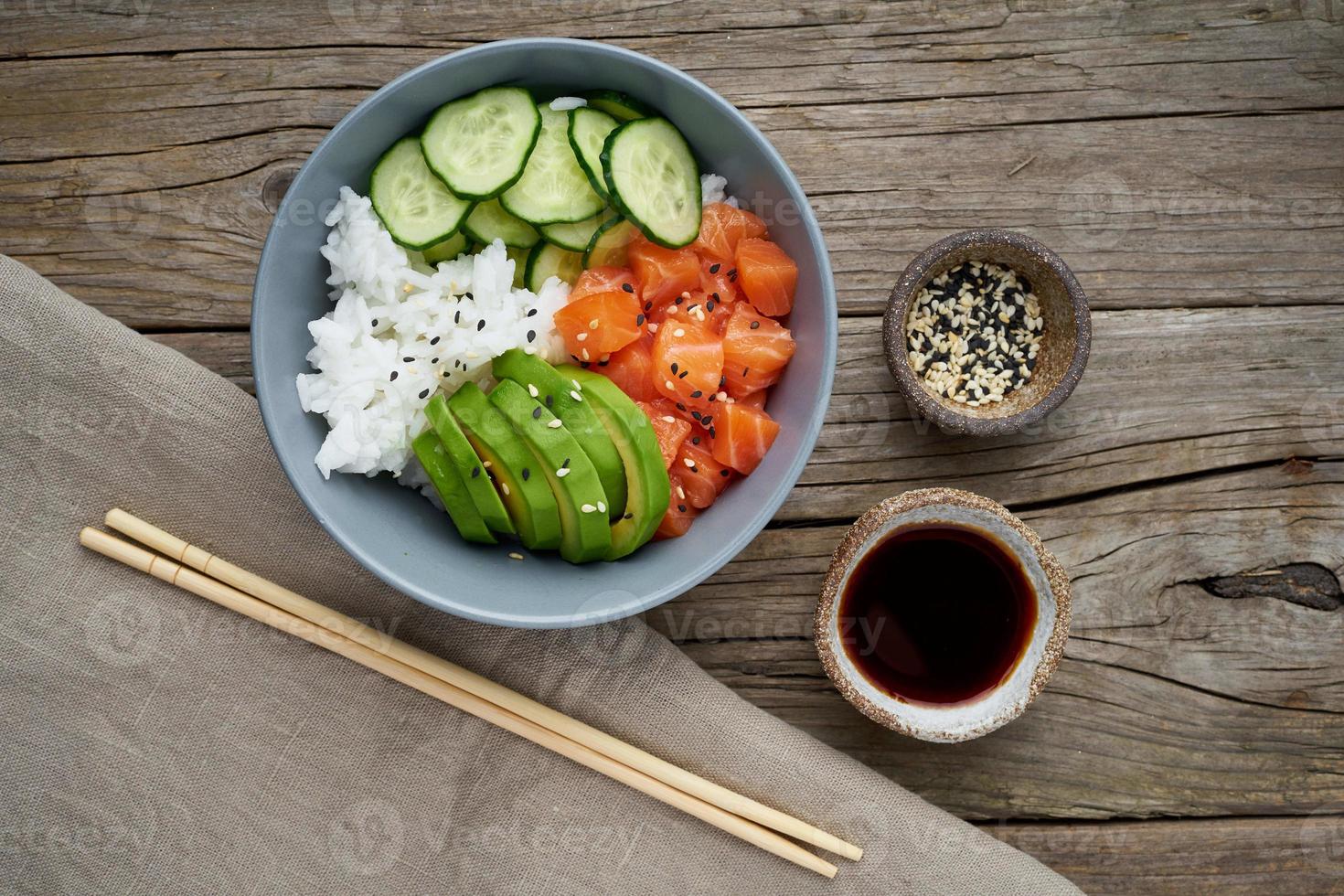 Salmon poke bowl with fresh fish, rice, cucumber, avocado with black and white sesame. Old wooden table. Food concept. photo