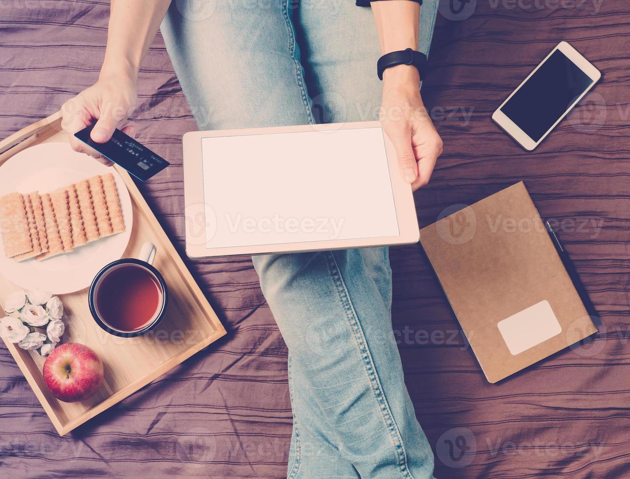 Woman sitting on bed, drinking tea and looking at tablet, pays for shopping, online shopping photo