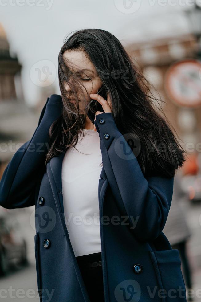 mujer con cabello largo hablando por teléfono. otoño o invierno, chica al aire libre. hermosa morena inteligente en la calle de san petersburgo en el centro de la ciudad, espacio de copia foto