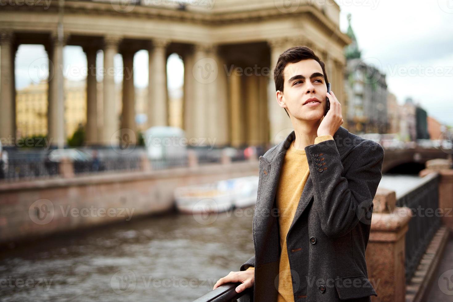 un apuesto hombre de moda está hablando por teléfono, una morena con un elegante abrigo gris está parada en la calle en el centro histórico de st. petersburgo joven de cabello oscuro, cejas pobladas. foto