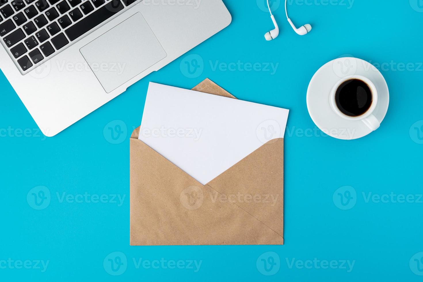 Open envelope with blank paper sheet on turquoise background. Keyboard, cup of coffee and earphones photo