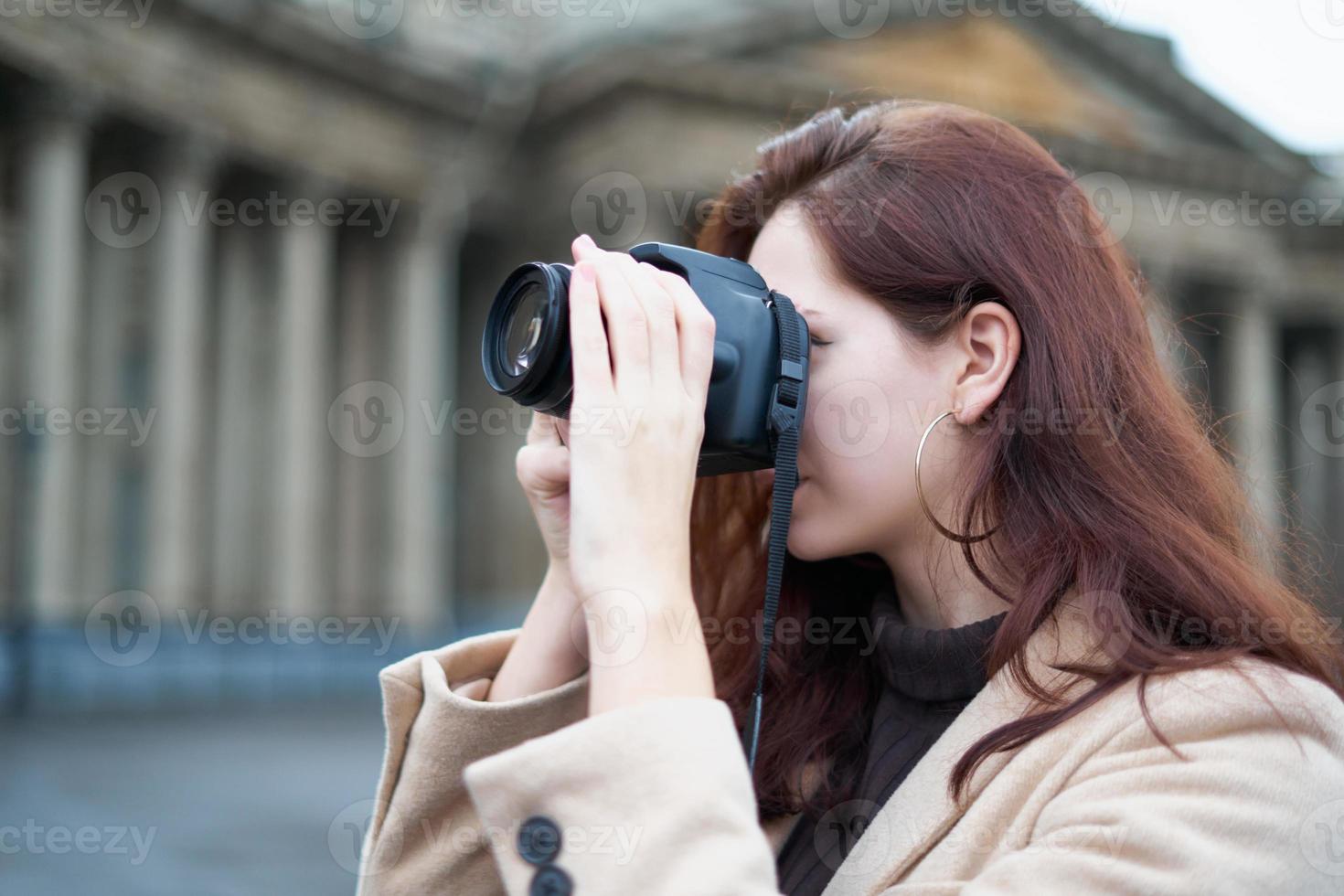 Beautiful stylish fashionable girl holds camera in her hands and takes pictures. Woman photographer with long dark hair in city, urban shoot. Unrecognizable person photo