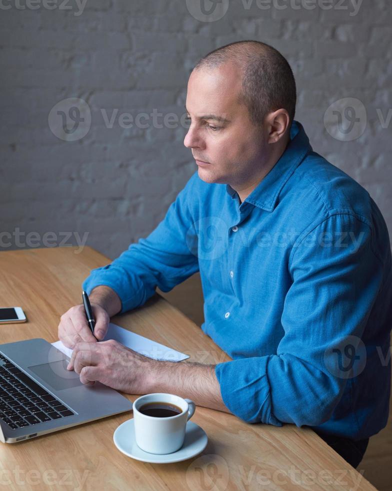 Handsome handsome mature man writes on a sheet of paper and looks at the computer screen, laptop. Man with casual clothes in a blue shirt at a table in the office in front of the window photo