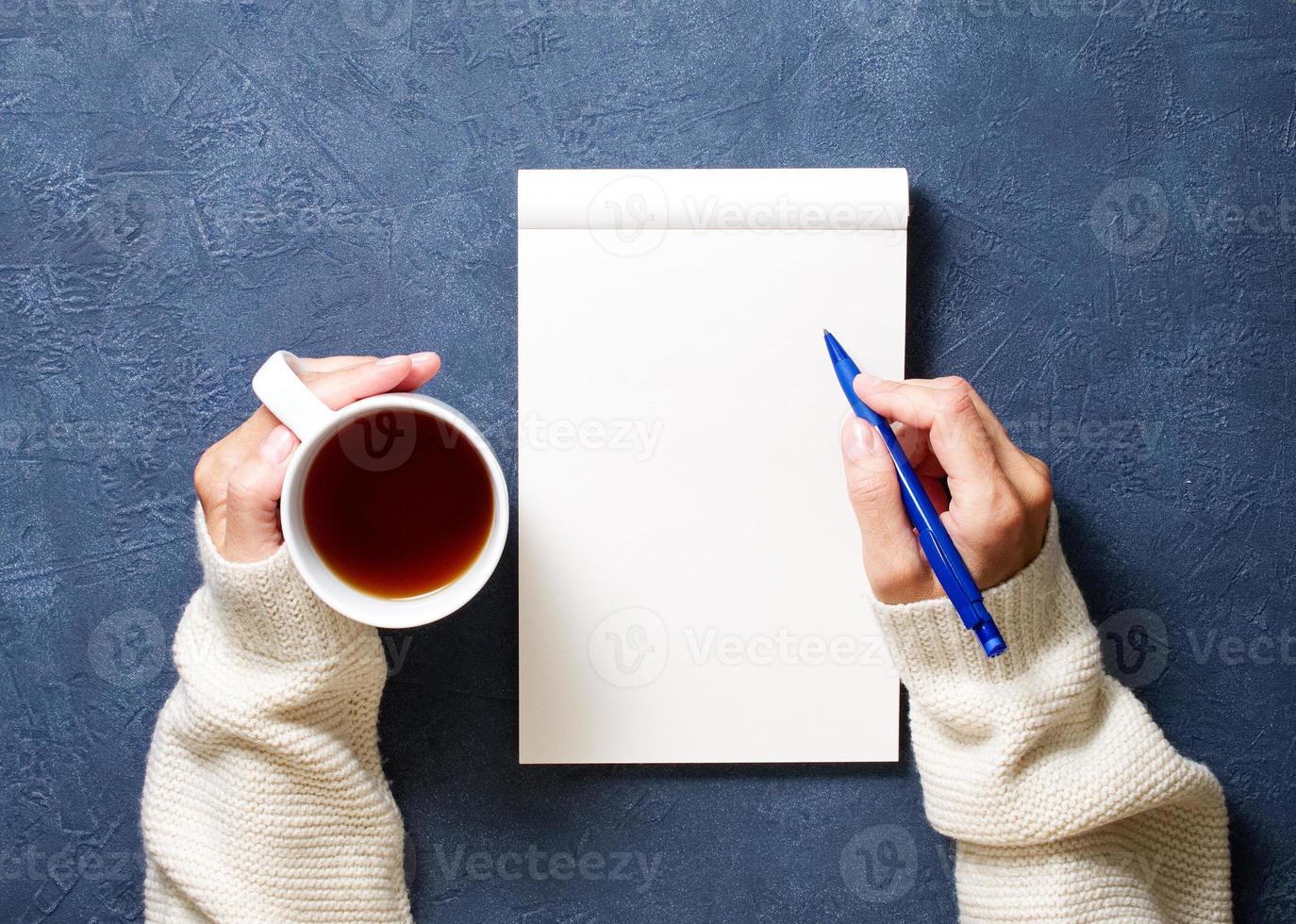 la mujer escribe en un cuaderno sobre una mesa azul oscuro, con la mano en el pantalones sosteniendo un lápiz, una taza de té, un dibujo de un cuaderno de bocetos, una vista superior foto