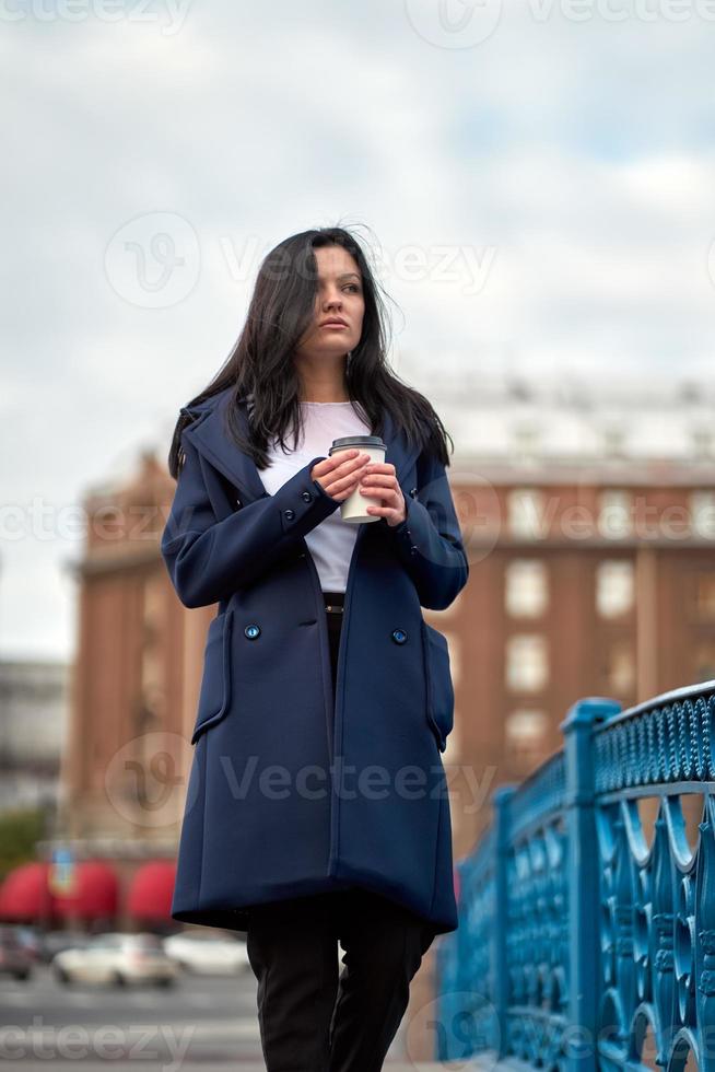 hermosa chica morena inteligente seria sosteniendo una taza de café en las manos va caminando por la calle de st. petersburgo en el centro de la ciudad en el puente. encantadora mujer pensativa con cabello largo y oscuro deambula sola foto