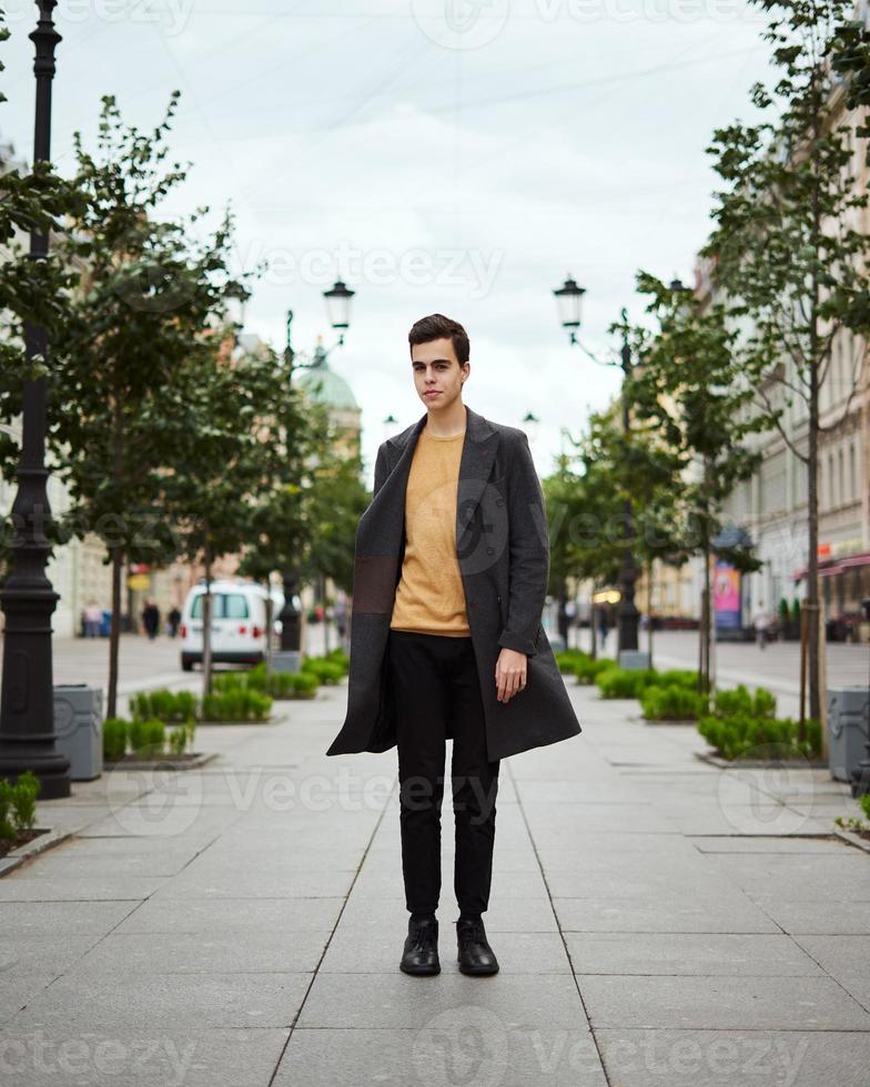 apuesto hombre de moda con estilo, morena con elegante abrigo gris, se encuentra en la calle en el centro histórico de st. petersburgo joven de cabello oscuro, cejas pobladas. foto