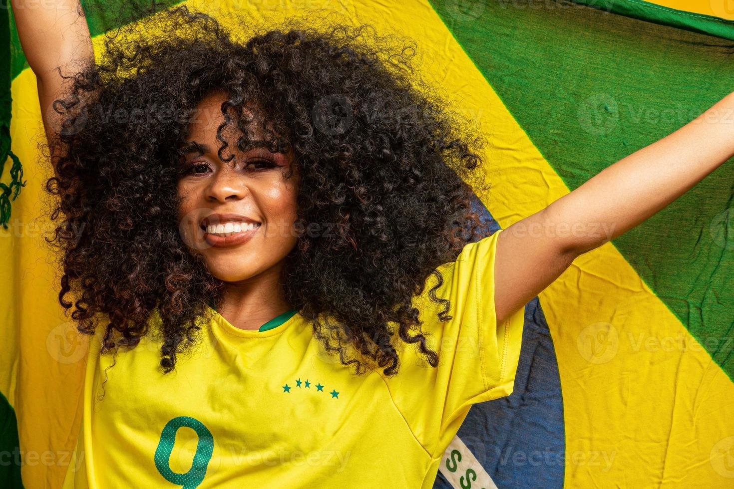 chica afro animando al equipo brasileño favorito, sosteniendo la bandera nacional en fondo amarillo. foto