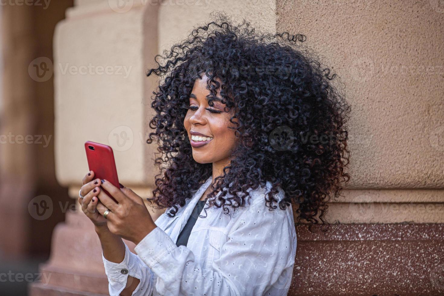 Young curly hair black woman walking using cell phone. Texting on street. Big city. photo