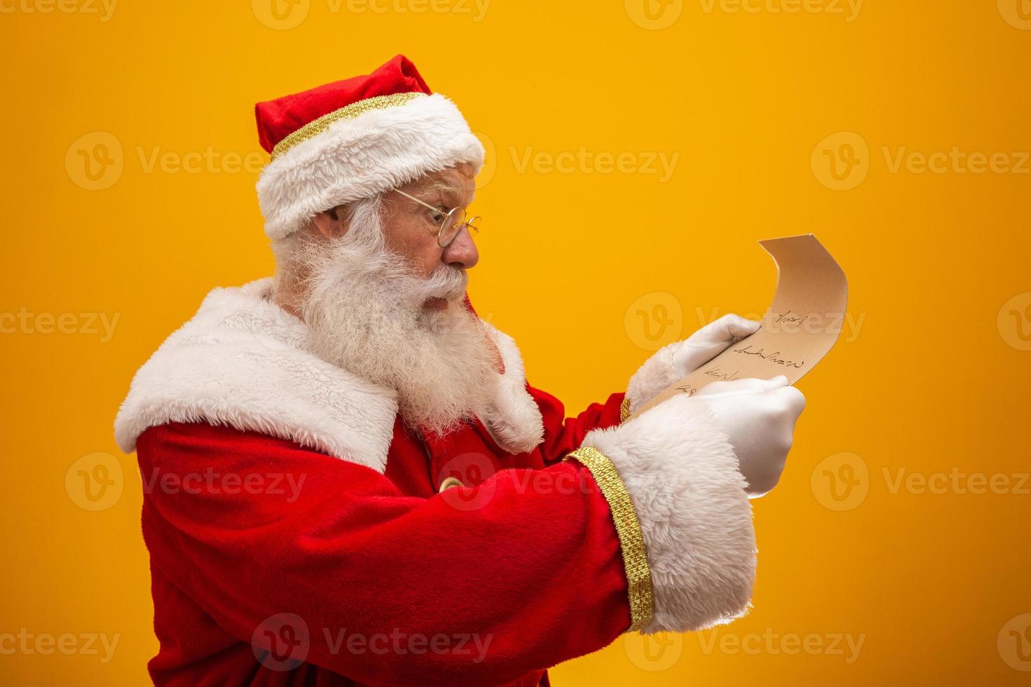 Happy Santa Claus holding vintage paper on yellow background. Merry Christmas and New Year's Eve concept. Copy space. Difficulty reading. Ugly handwriting. photo