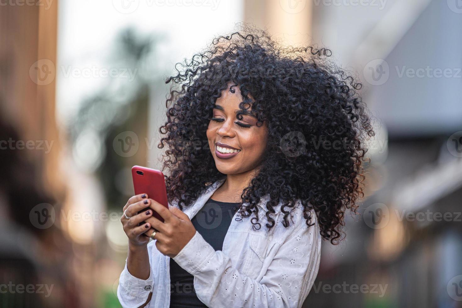 joven mujer negra de pelo rizado caminando con teléfono celular. enviando mensajes de texto en la calle. Gran ciudad. foto