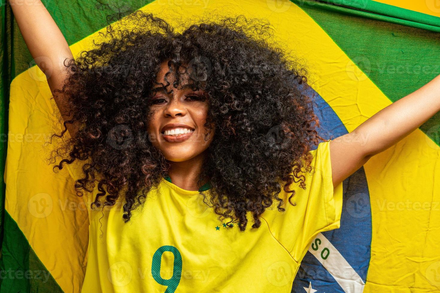chica afro animando al equipo brasileño favorito, sosteniendo la bandera nacional en fondo amarillo. foto