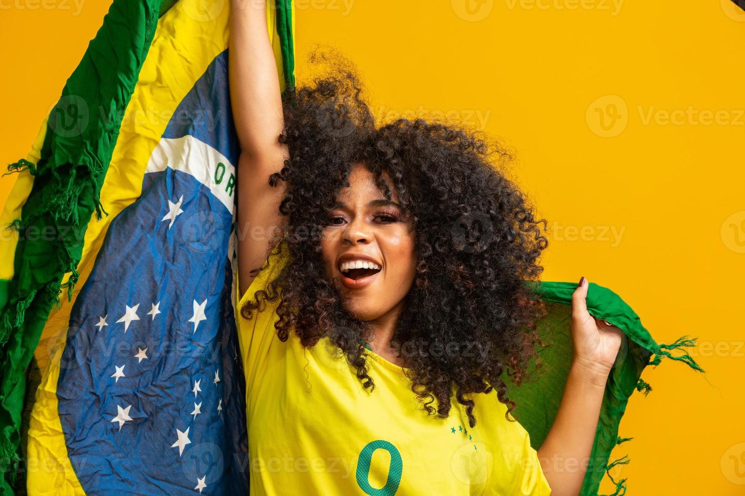 chica afro animando al equipo brasileño favorito, sosteniendo la bandera nacional en fondo amarillo. foto