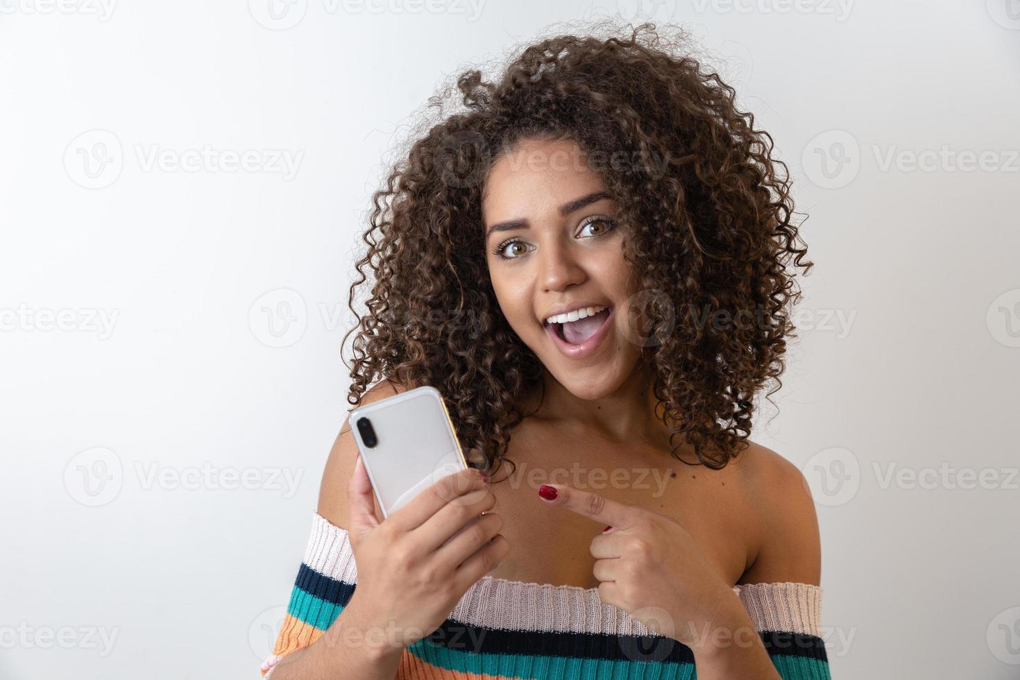 Studio shot of entertained cute happy african american girl with afro hairstyle holding smartphone using device to have fun photo