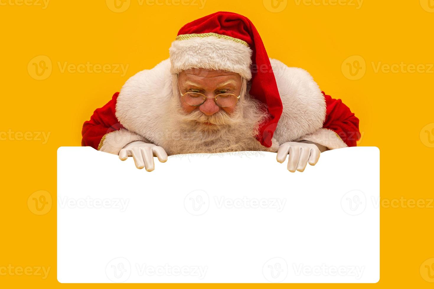 Happy Santa Claus looking out from behind the blank sign isolated on yellow background with copy space photo