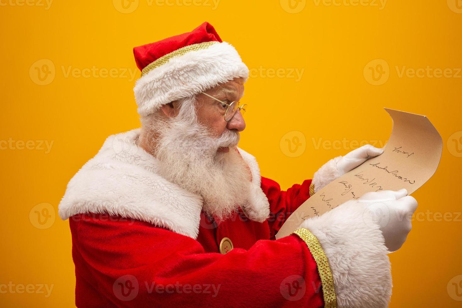 Happy Santa Claus holding vintage paper on yellow background. Merry Christmas and New Year's Eve concept. Copy space. Difficulty reading. Ugly handwriting. photo