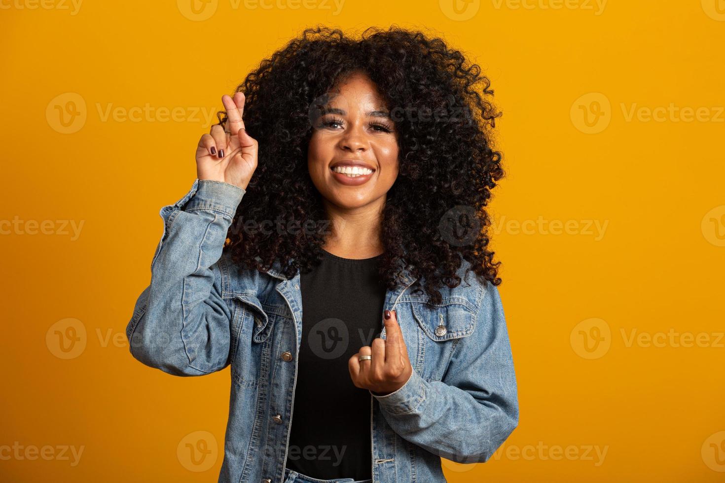 I have to win. Joyful young brunette female clenches teeth, raises fingers crossed, makes desirable wish, curly hair, mixed race, African, Brazilian girl, waits for good news, stands over yellow wall photo