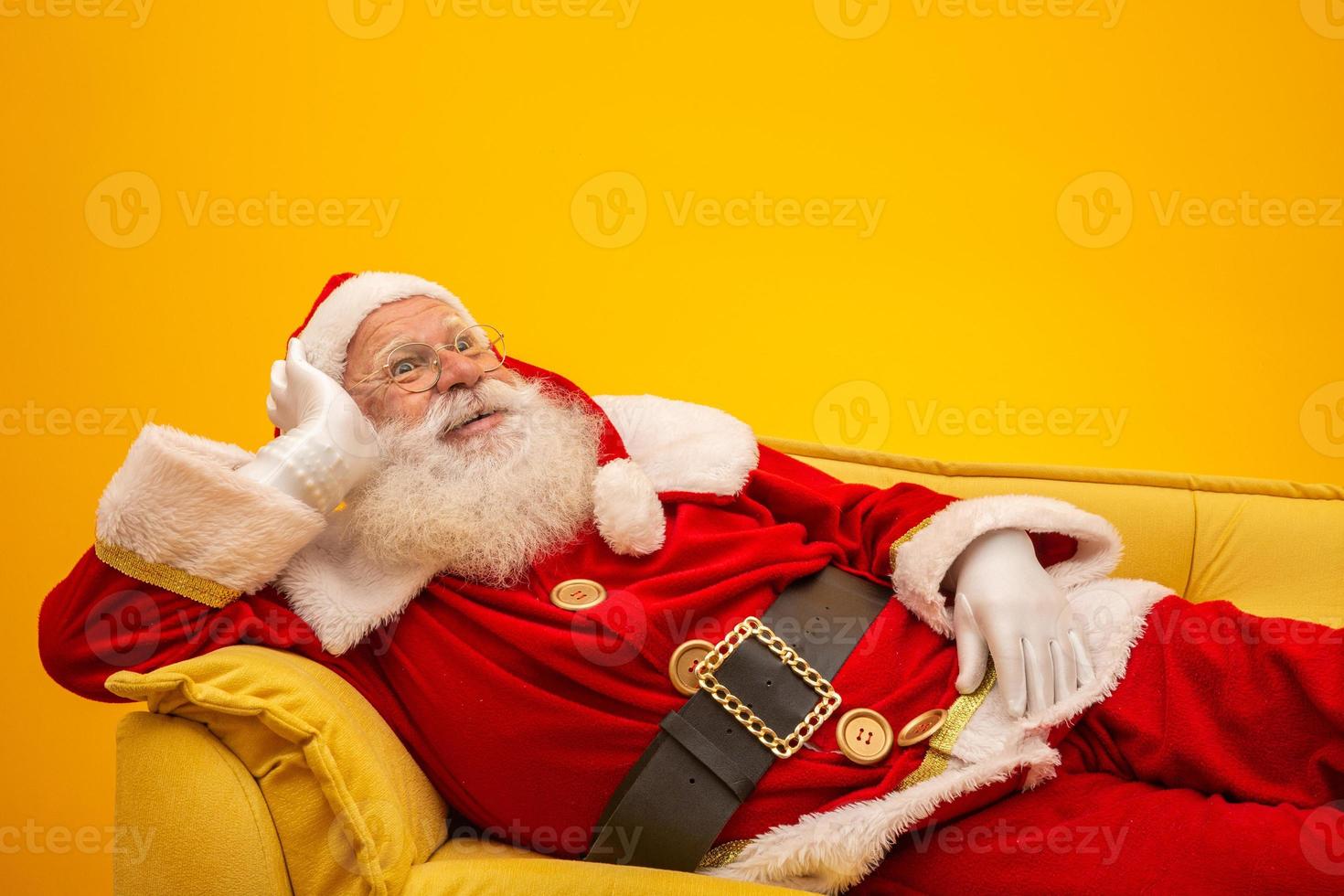 Santa Claus sitting on a yellow couch on yellow background with copy space. Yellow sofa. photo