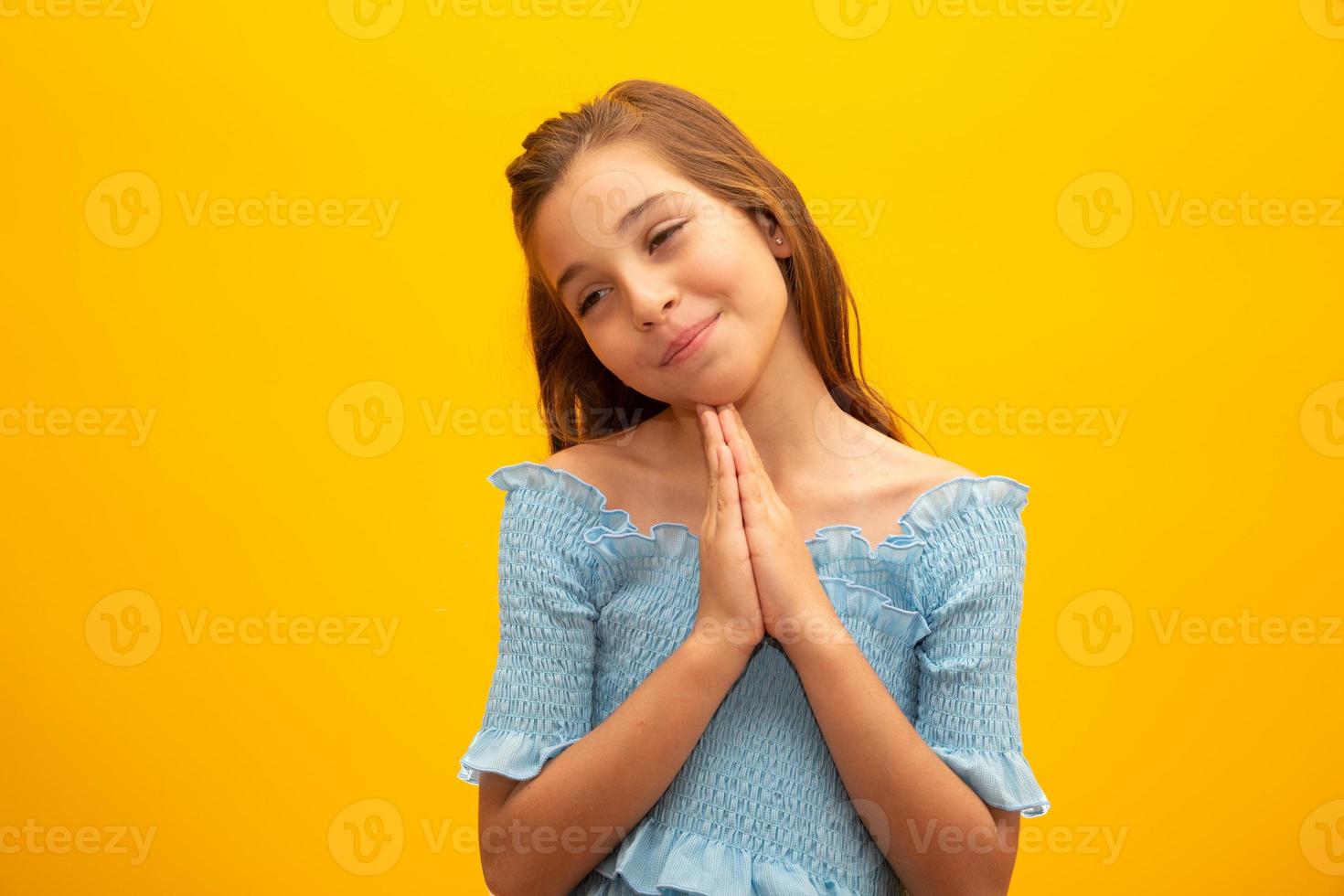 Nice little girl, holds hands in praying gesture, stands over isolated yellow background with copy space. photo