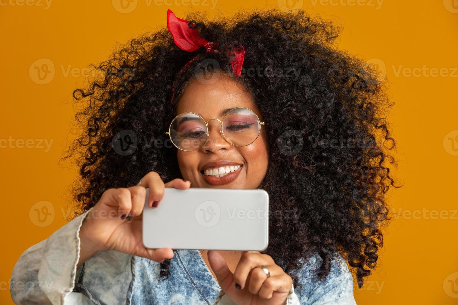 Studio shot of entertained cute happy african american girl with afro hairstyle holding smartphone using device to have fun. Yellow background. photo