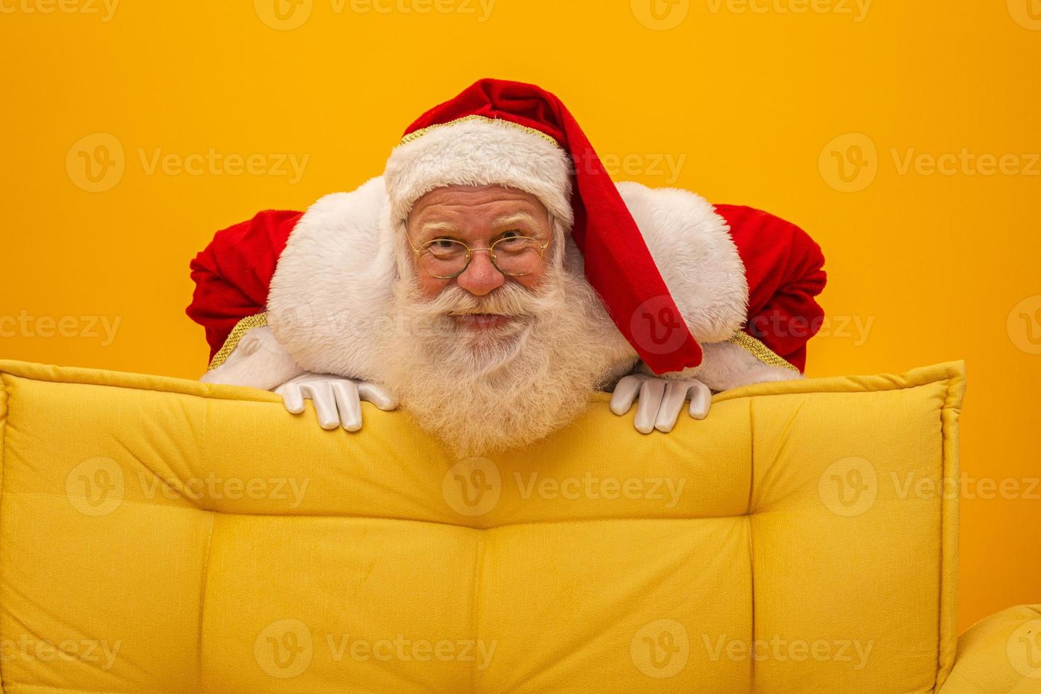 Santa Claus sitting on a yellow couch on yellow background with copy space. Yellow sofa. photo