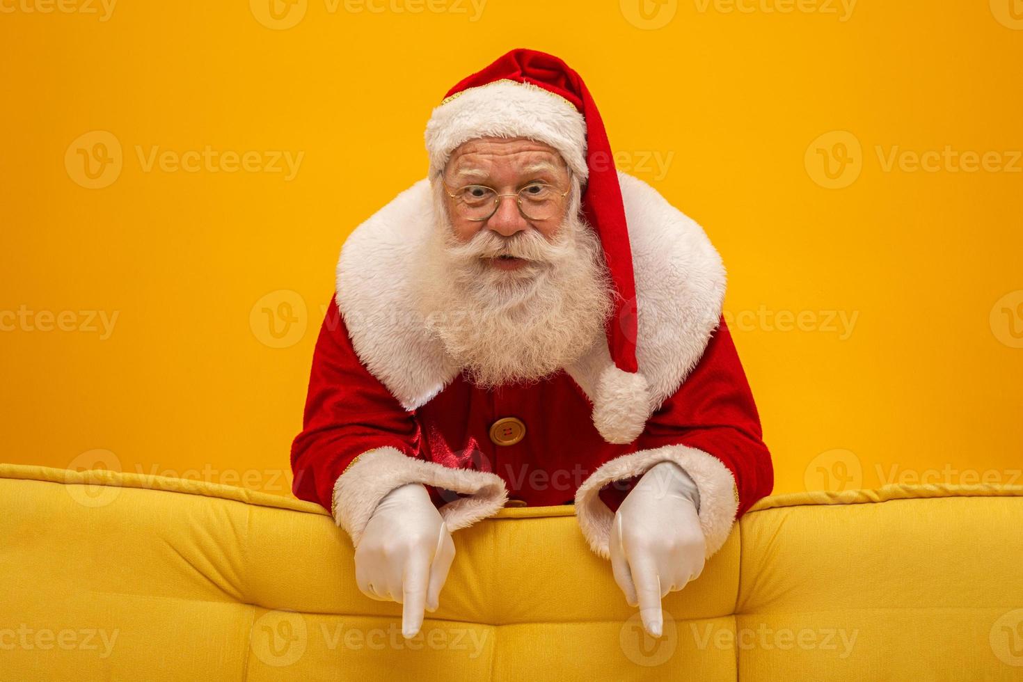 Santa Claus sitting on a yellow couch on yellow background with copy space. Yellow sofa. Pointing down. photo