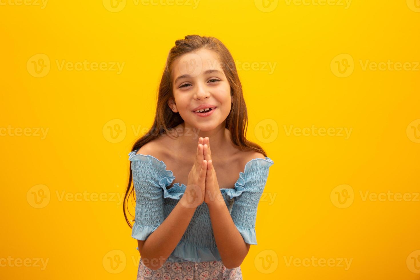Nice little girl, holds hands in praying gesture, stands over isolated yellow background with copy space. photo