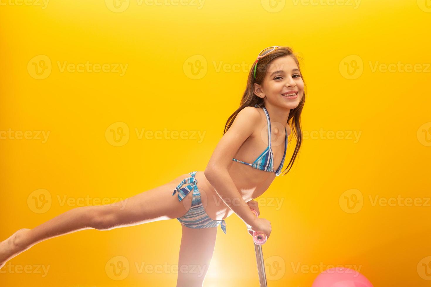 Child in bikini on yellow background playing with his scooter. photo