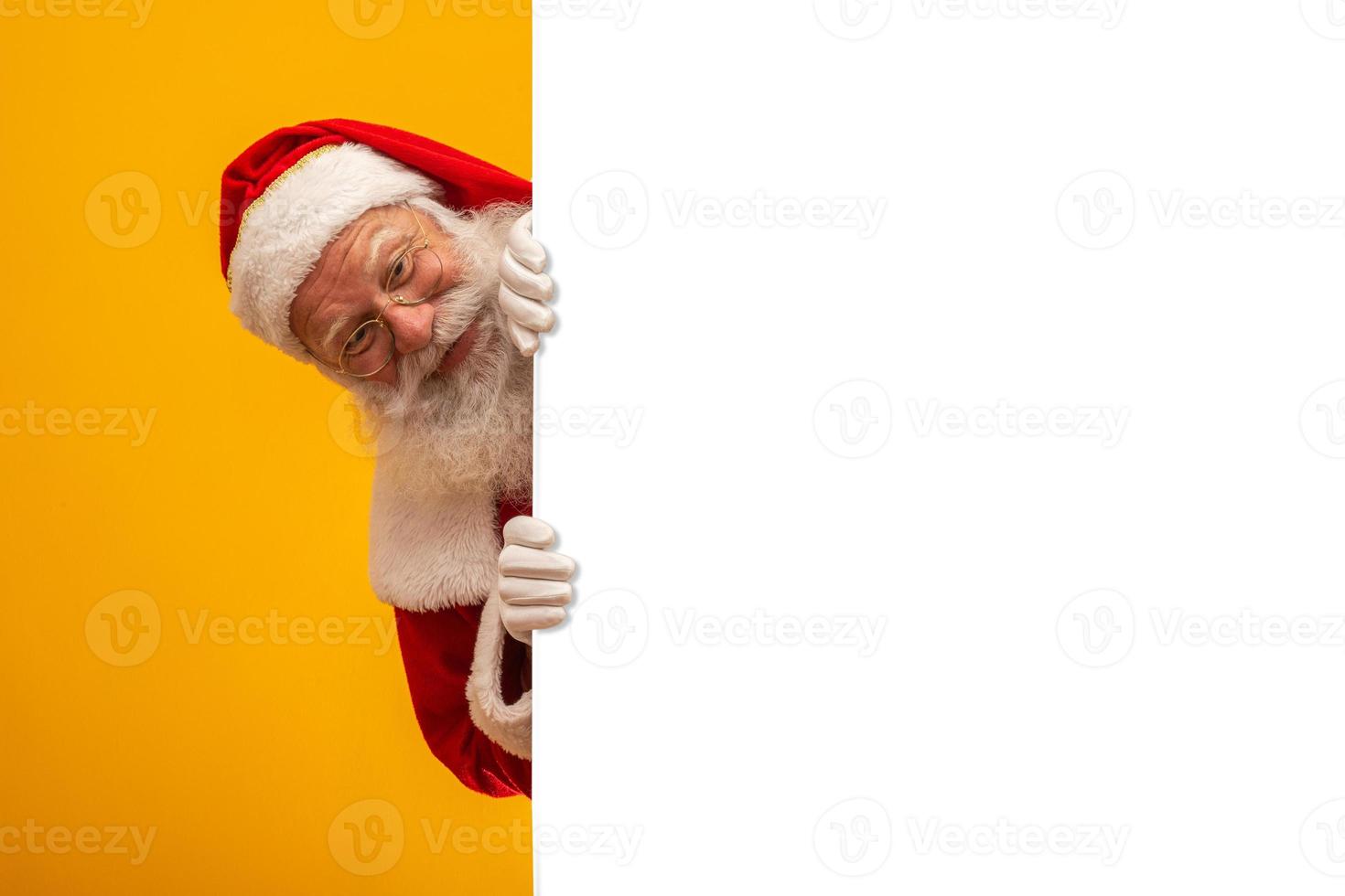 Happy Santa Claus looking out from behind the blank sign isolated on yellow background with copy space photo