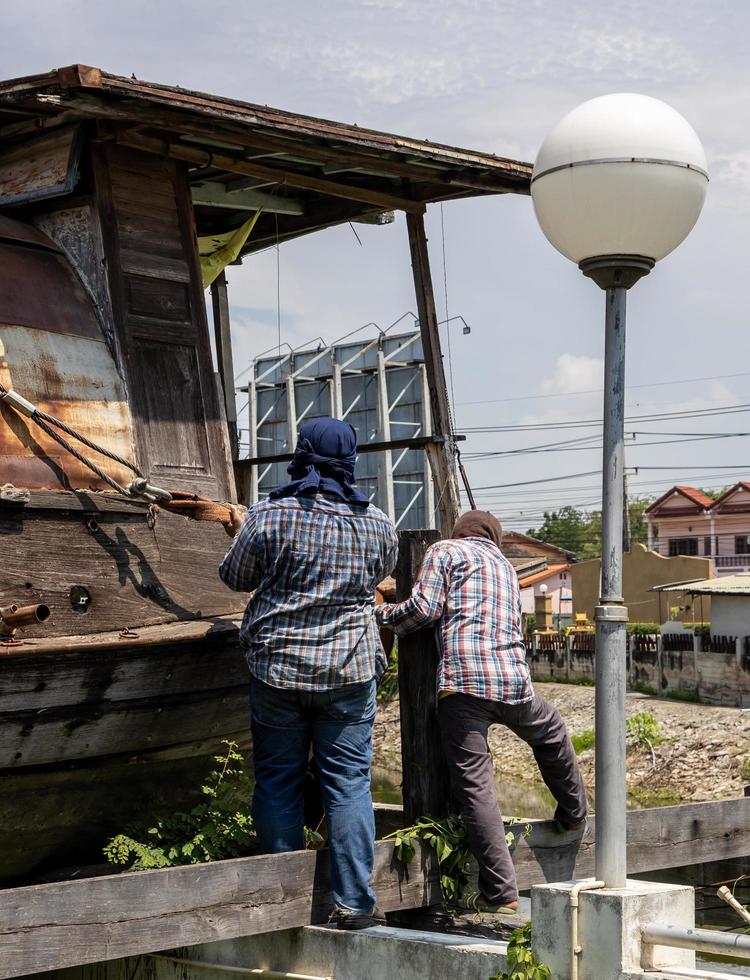 un grupo de trabajadores tailandeses está instalando grandes eslingas de cuerda para levantar y mover un viejo bote de madera. foto