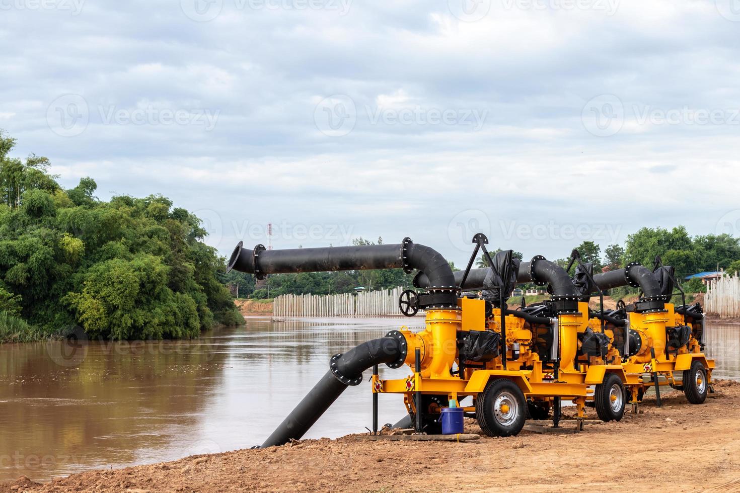 una gran bomba amarilla con tuberías negras está montada en el suelo de la orilla del río en preparación para bombear cerca de una presa, una pared de columna de hormigón para evitar la erosión. foto