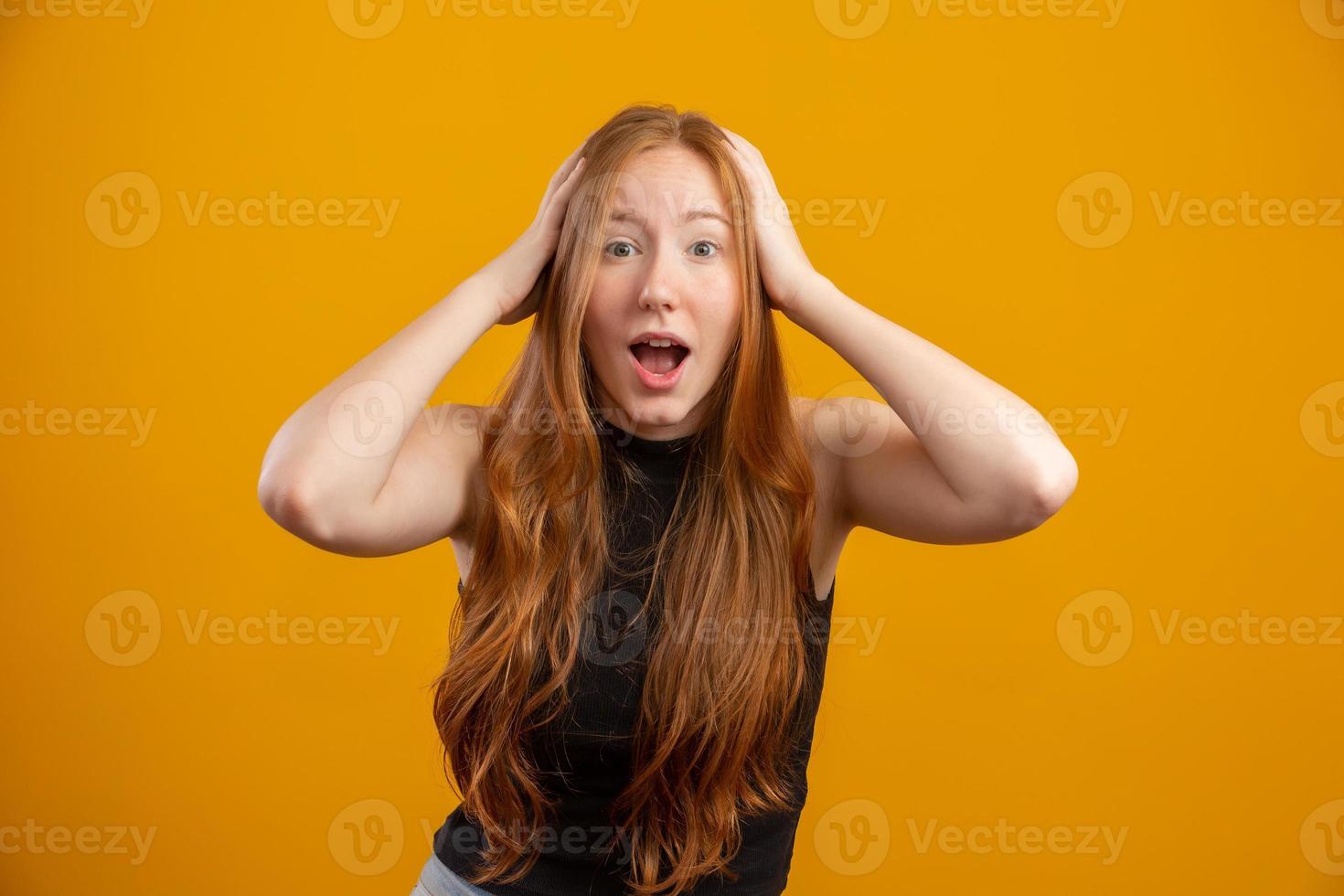 joven pelirroja levantando las manos a la cabeza, con la boca abierta, sintiéndose extremadamente afortunada, sorprendida, emocionada y feliz contra la pared amarilla. foto