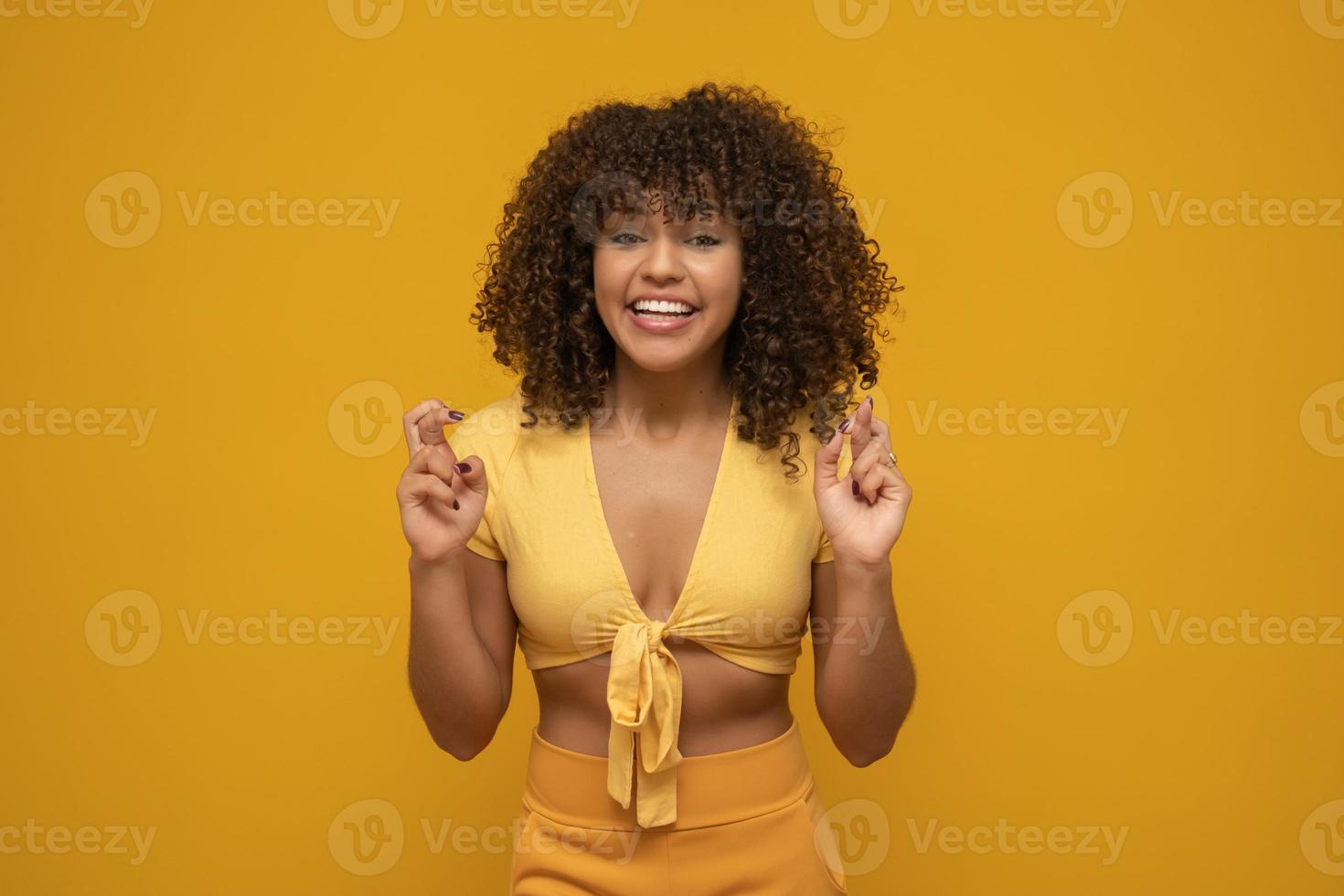 retrato de una joven sonriente con el pelo largo y rizado de morena sobre un fondo amarillo, con los dedos cruzados para la buena suerte. foto