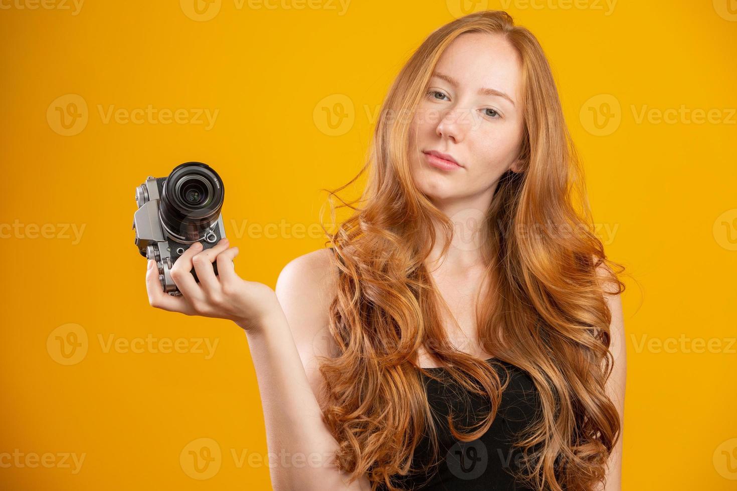 Photo of adorable redhead woman wearing black clothes holding retro vintage camera and taking picture isolated over yellow background. Mock up copy space. Hold retro vintage photo camera.