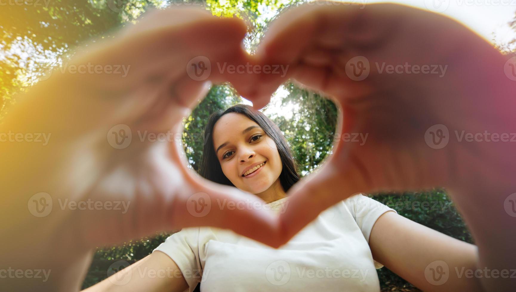 Cute happy teenager girl making heart shape hand gesture looking at camera, funny smiling ethnic young single woman blogger laughing face showing love sign symbol, park, close up portrait photo