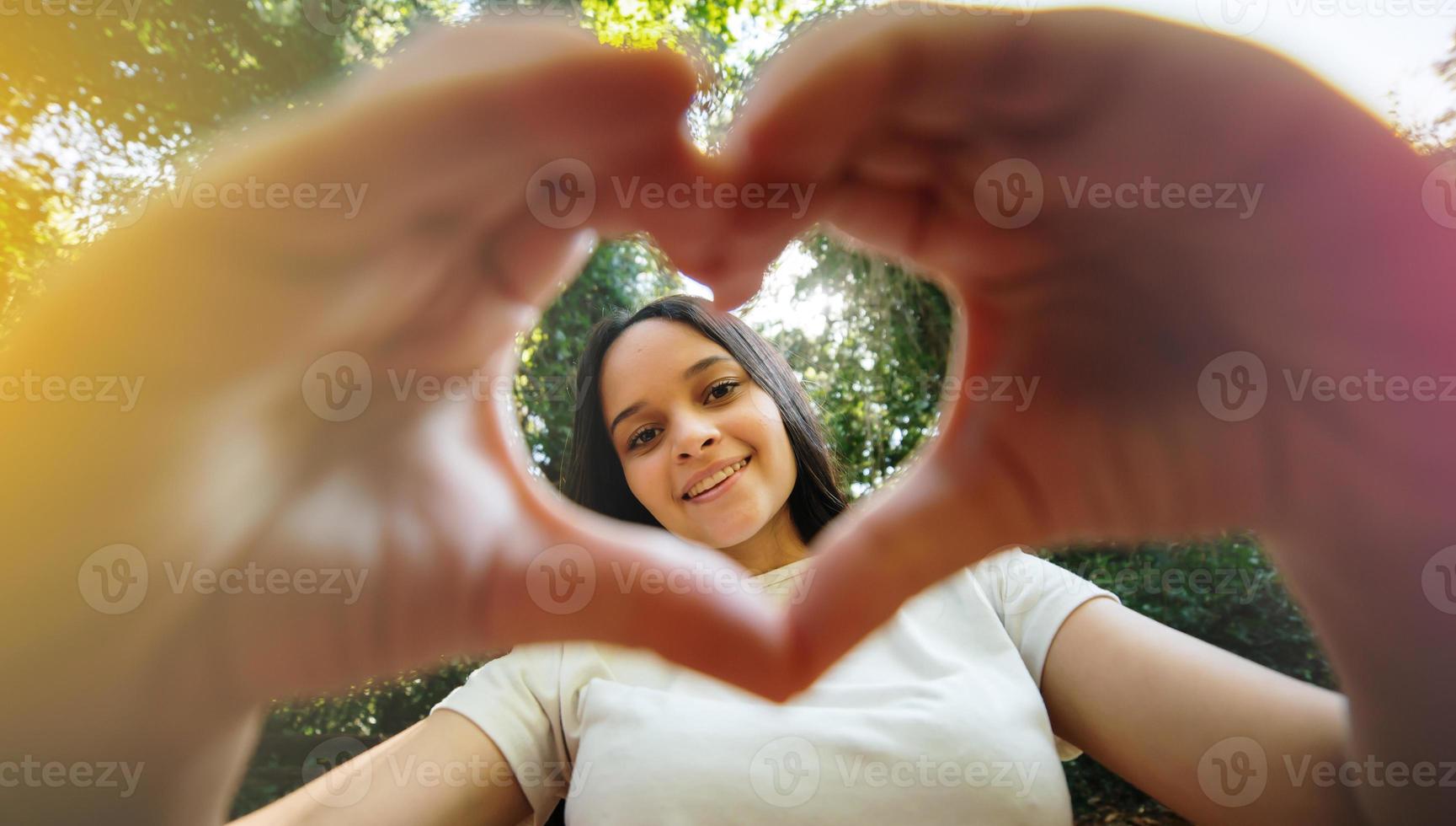 linda adolescente feliz haciendo gestos con la mano en forma de corazón mirando a la cámara, graciosa sonriente étnica joven bloguera soltera riéndose cara mostrando símbolo de signo de amor, parque, retrato de cerca foto