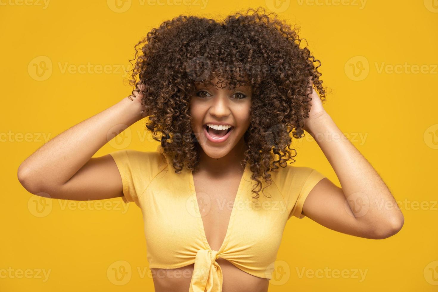 feliz risa mujer africana americana con su pelo rizado sobre fondo amarillo. mujer rizada riendo en suéter tocando su cabello y mirando a la cámara. foto