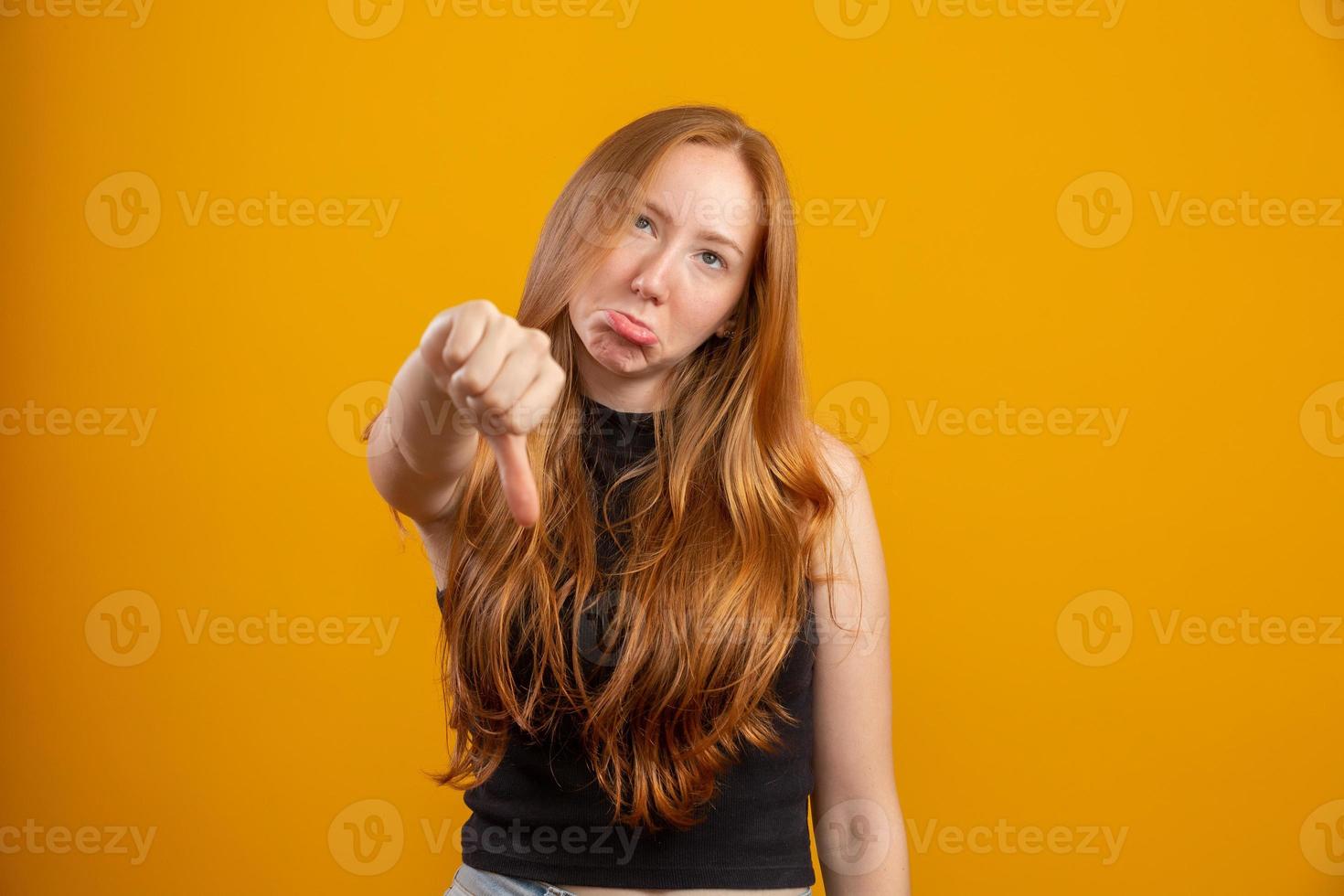 Unhappy redhead woman giving thumbs down gesture looking with negative expression and disapproval. Yellow background. photo