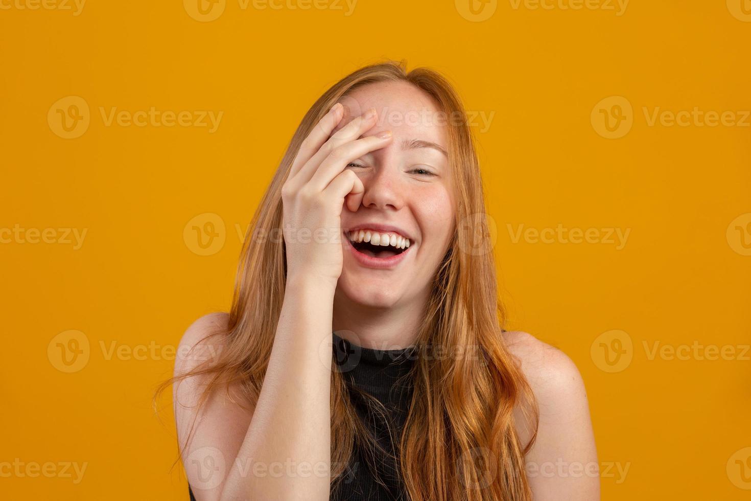 Una joven asustada de cabello rojo aislada en un estudio amarillo se esconde detrás de las manos, una pelirroja aterrorizada se cubre la cara mirando a través de los dedos, una mujer curiosa siente miedo de mirar a escondidas. foto