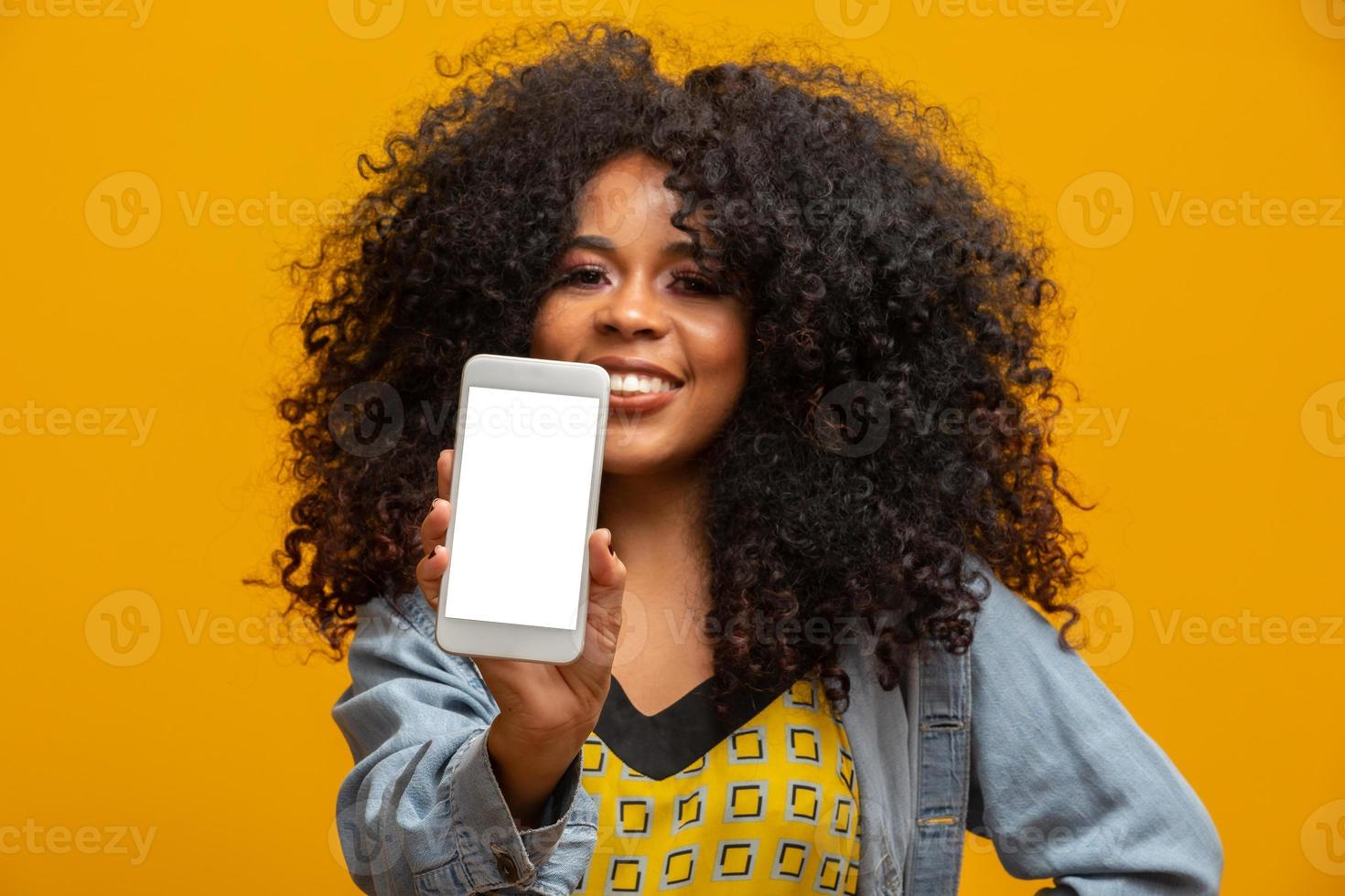 Portrait of cheerful, positive, attractive young woman in jeans shirt, having smart phone with white screen in hand, pointing with forefinger to product, isolated on yellow background photo