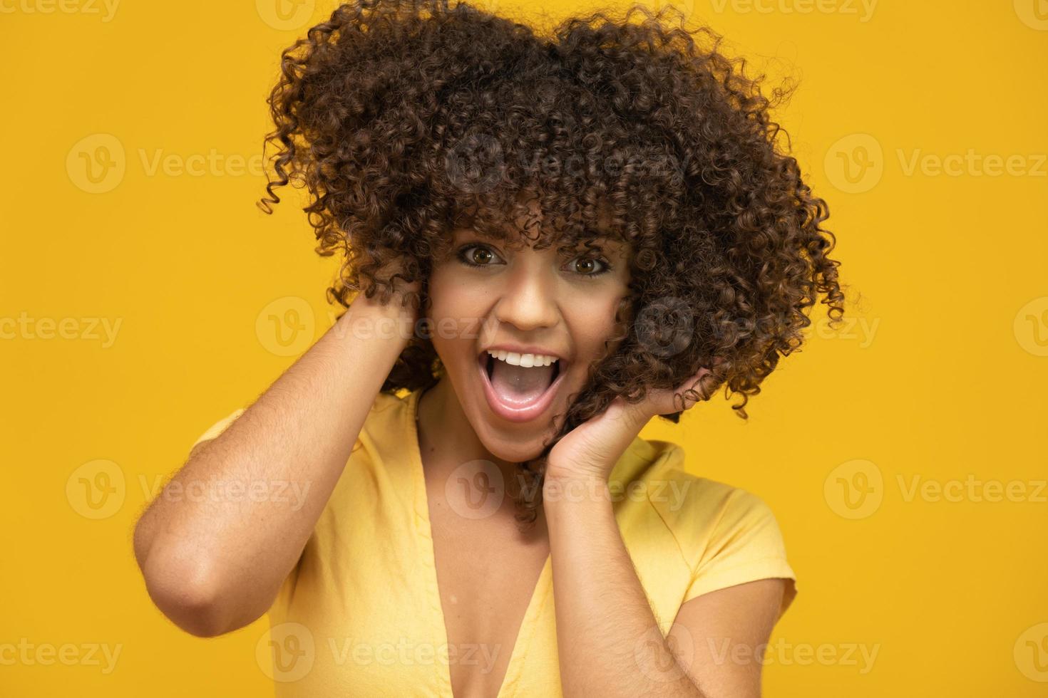 feliz risa mujer africana americana con su pelo rizado sobre fondo amarillo. mujer rizada riendo en suéter tocando su cabello y mirando a la cámara. foto