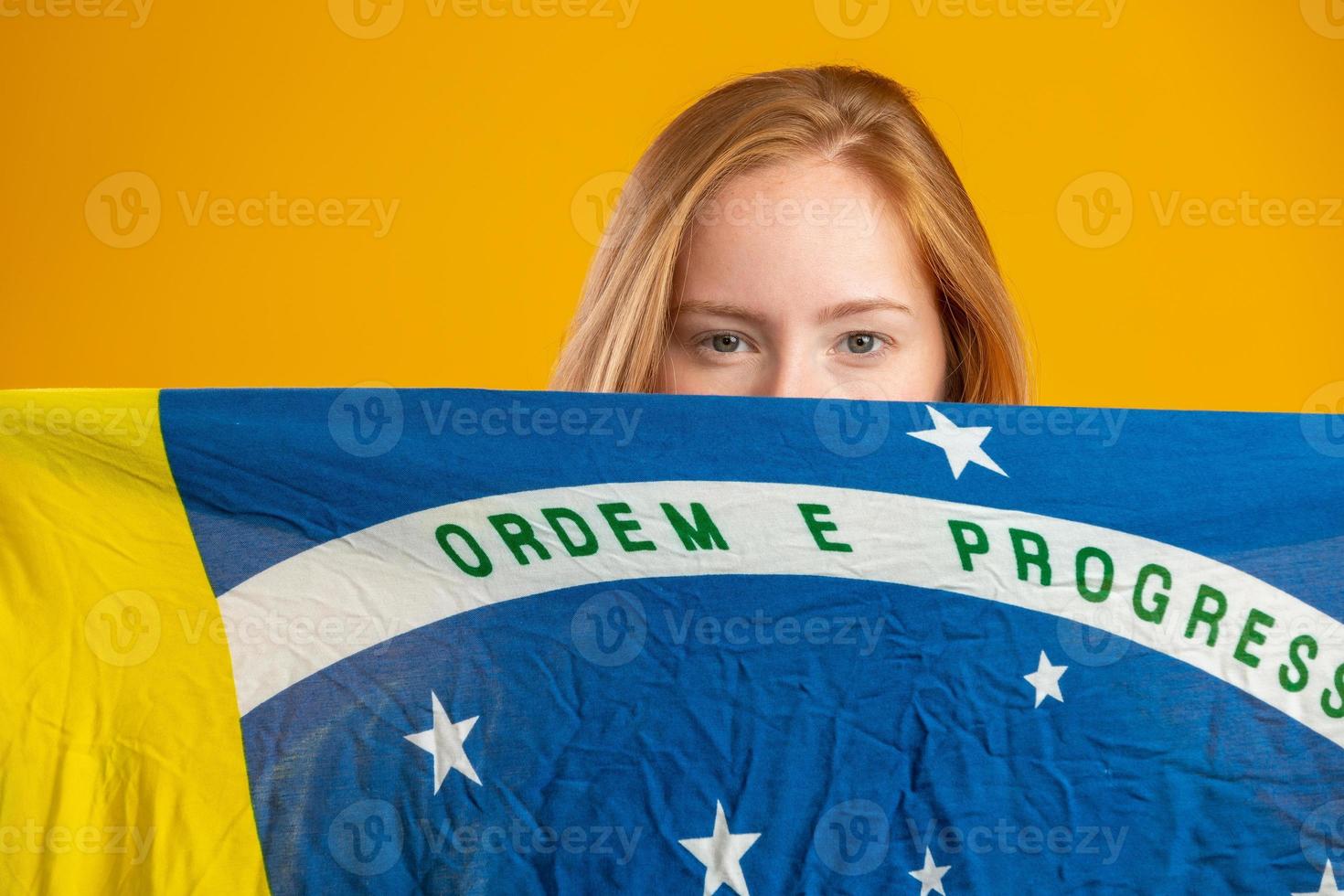 misteriosa fan de mujer pelirroja sosteniendo una bandera brasileña en tu cara. colores brasil de fondo, verde, azul y amarillo. elecciones, fútbol o política. foto