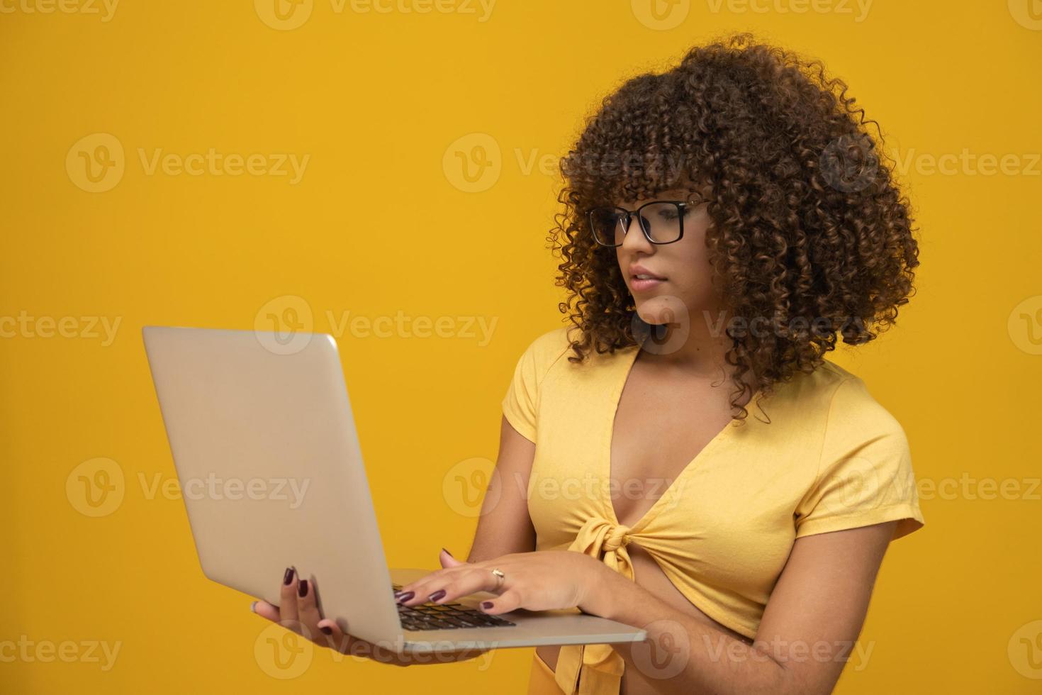 retrato de una sonriente joven de raza mixta de pelo rizado sosteniendo un ordenador portátil aislado sobre fondo amarillo. foto