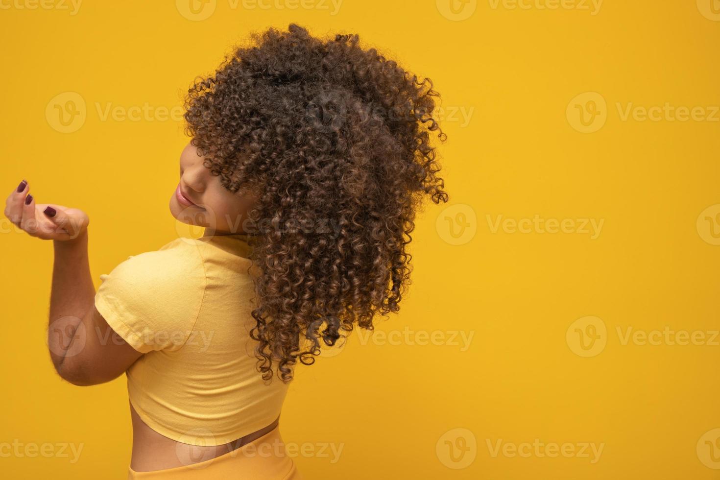 Backwards American African woman with her curly hair on yellow background. Laughing curly woman in sweater touching her hair and looking at the camera. photo