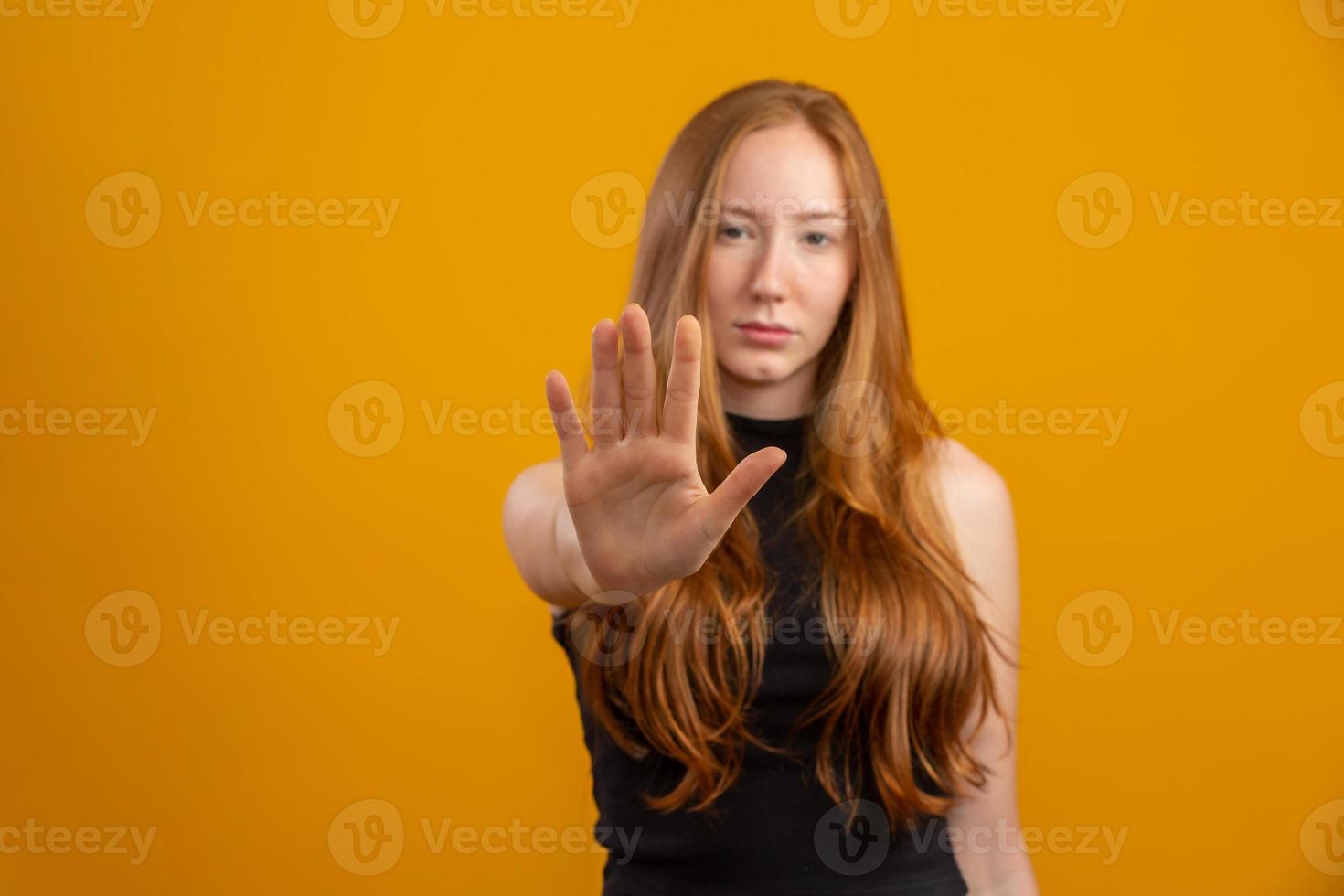 Beautiful redhead woman standing over isolated yellow background with open hand doing stop sign with serious and confident expression, defense gesture. No more violence against women. Abuse. photo