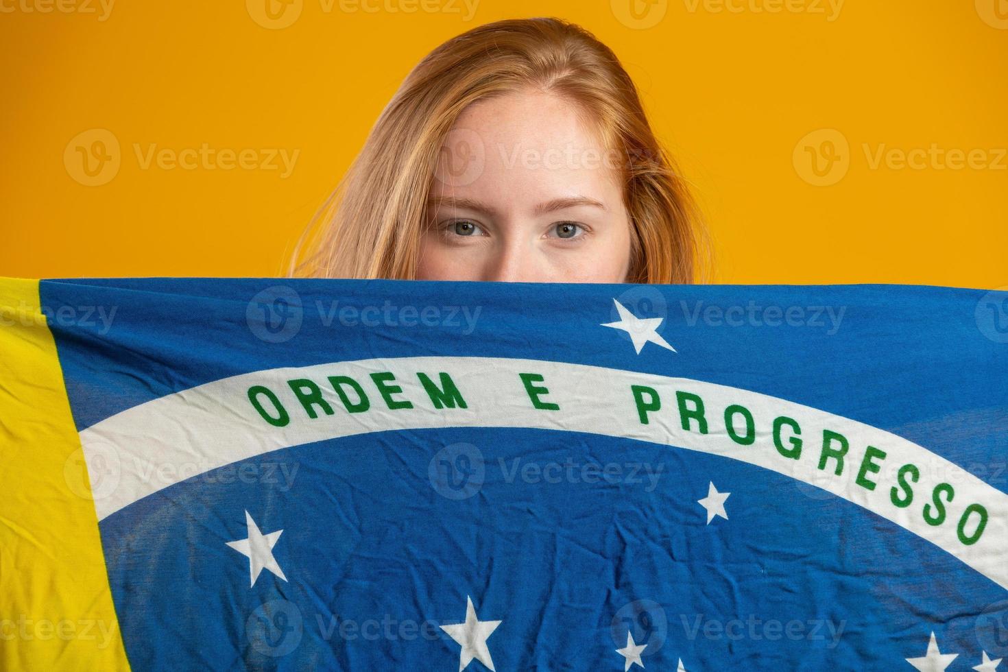 misteriosa fan de mujer pelirroja sosteniendo una bandera brasileña en tu cara. colores brasil de fondo, verde, azul y amarillo. elecciones, fútbol o política. foto