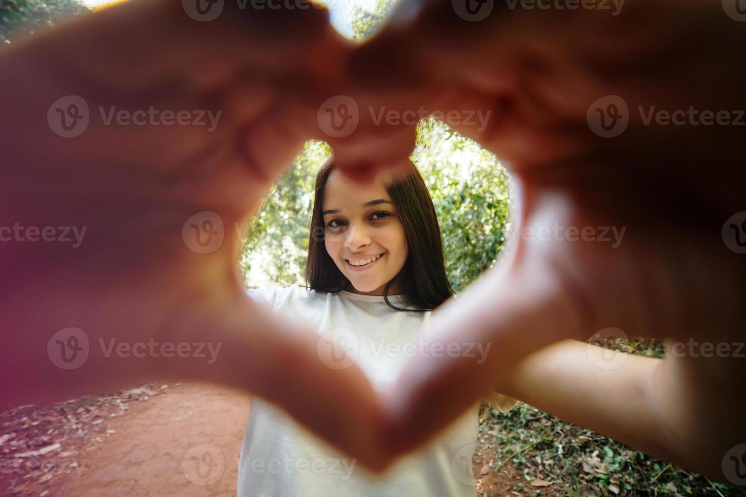 Cute happy teenager girl making heart shape hand gesture looking at camera, funny smiling ethnic young single woman blogger laughing face showing love sign symbol, park, close up portrait photo